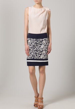 Haut sans manches crème avec des détails plissés, associé à une jupe crayon à motifs floraux navy avec des accents à rayures blanches. Sandales à talons bruns.