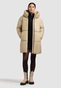 Abrigo acolchado beige con capucha, que presenta una cremallera frontal, diseño acolchado y bolsillos laterales. El modelo lleva un vestido negro de canalé y botas de tobillo beige.