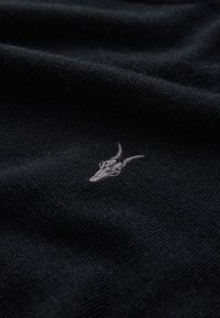 Schwarzer gestrickter Stoff mit einem kleinen grauen bestickten Detaildesign eines Widderkopfes. Die Textur ist glatt mit einem gleichmäßigen Gewebe.