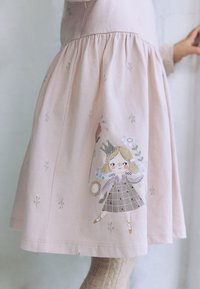 Ružové bavlnené šaty so skladanou sukňou. Obsahujú vyšívaný motív víly a kvetinové vzory, s jemnou, hladkou textúrou.