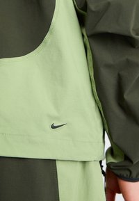 Žalia ir tamsiai žalia trumpa palaidinė su apvaliu iškirpimu, ant kurios yra juodas Nike logotipas. Medžiaga atrodo lygi, matomi siuvimo detalės.