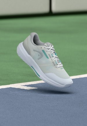 Lichtgrijze sportschoen met een mesh bovenwerk met turquoise accenten, een gevoerde zool en gedetailleerde veters, geplaatst bovenop een blauw sportveld.