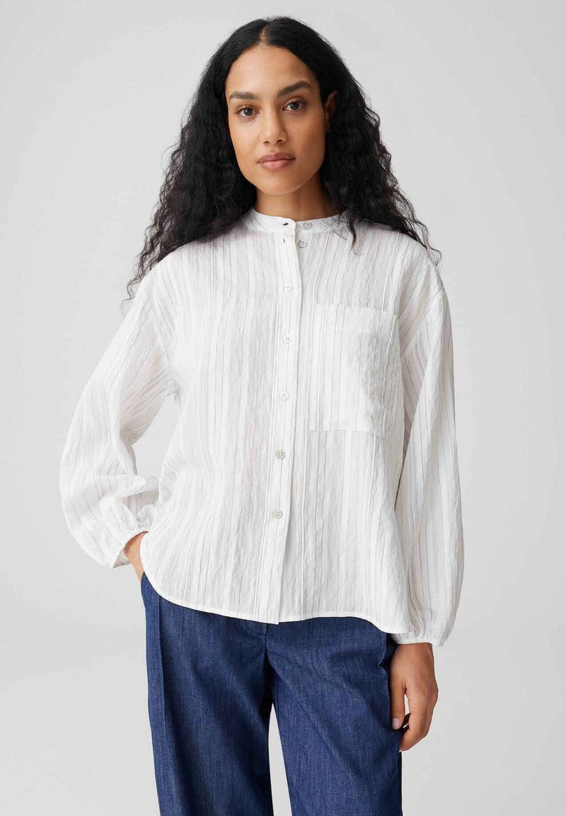 Vrouw met lang, krullend haar die een witte blouse met knopen en gestructureerde strepen en blauwe broek draagt, staand met haar hand in haar zak.