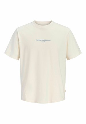 Blond chłopiec stojący, ubrany w kremowy krótki rękaw T-shirt, ciemne szerokie jeansowe szorty, białe skarpetki i brązowe sznurowane buty na jednolitym tle.