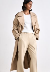 Manteau beige en tissu à motif, devant croisé et larges revers, associé à un haut crème et un pantalon beige.