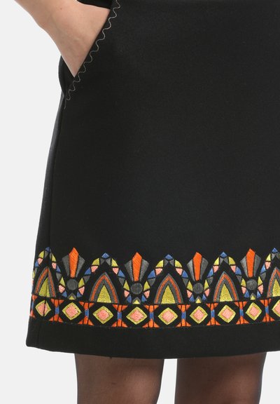 Crna suknja s složenim višebojnim vezenjem na rubu. Sadrži geometrijske uzorke u narančastoj, žutoj, ružičastoj i plavoj boji. Obod džepa je valovit.