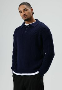 Jonge man met gevlochten haar, gekleed in een donkerblauwe gebreide trui over een wit shirt, staand met handen in de zakken tegen een effen achtergrond.