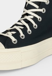Κοντινή λήψη ενός μαύρου sneaker ψηλού τύπου με λευκά κορδόνια, μεταλλικούς οφθαλμούς και παχιά λευκή καουτσούκ σόλα.