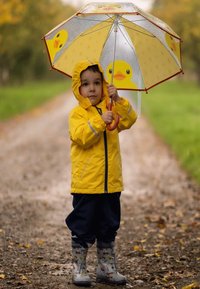 Geel regenjasje met reflecterende accenten, blauwe broek en grijs gestreepte regenlaarzen. Houdt een doorzichtige paraplu met gele eendenontwerpen.