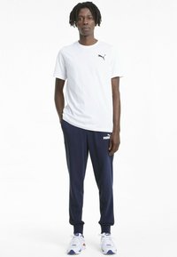 Bela majica s kratkimi rokavi z črnim logotipom Puma, v kombinaciji z mornarsko modrimi trenirkami, ki nosijo bel logotip Puma, in belimi superge z modrimi akcenti.