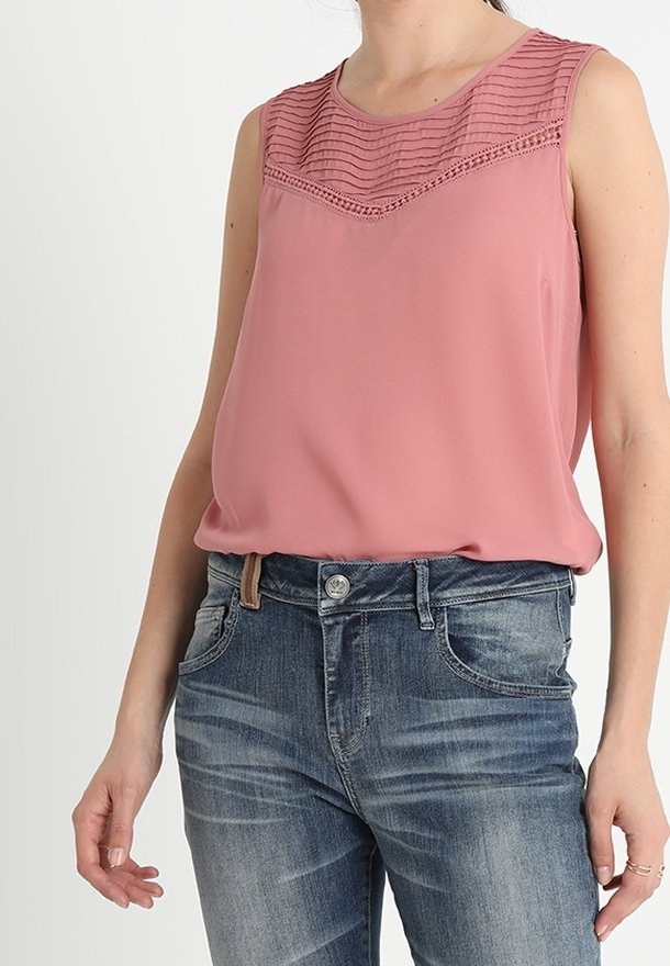 Femme portant un chemisier rose sans manches avec un empiècement texturé, rentré dans un jean bleu délavé avec poches avant et passants pour ceinture.
