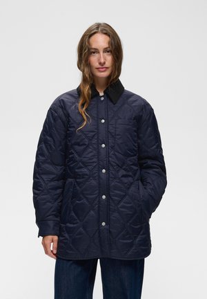 Frau mit langen welligen Haaren, die eine marineblaue gesteppte Jacke mit Knöpfen und schwarzem Kragen sowie dunkelblaue Jeans vor einem schlichten Hintergrund trägt.