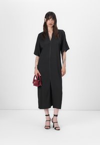 Black zip-front dress with short sleeves and a relaxed fit. Model holds a small red handbag; wears strappy black sandals.
