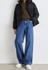 Doudoune noire, pull vert olive, jean bleu à jambes larges, baskets blanches. Caractéristiques : fermeture éclair et manches retroussées.