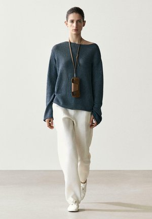 Kobieta spacerująca w białych spodniach, luźnym granatowym swetrze, białych butach oraz z brązową skórzaną torbą zawieszoną na długim pasku na szyi.