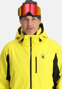 Helle gelbe Ski-Jacke mit schwarzen Akzenten, verstellbarer Kapuze und Reißverschlusstaschen. Ausgestattet mit rot getönten Skibrillen und einer grauen Mütze.