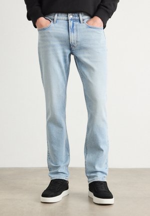 Muž nosící světle modré džíny rovného střihu s rukama v kapsách a černé tenisky s bílými podrážkami, stojící na šedé podlaze.