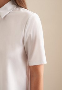 Camicetta bianca a maniche corte con colletto, realizzata in tessuto morbido e con una vestibilità rilassata. Cuciture visibili e un design pulito e minimale.