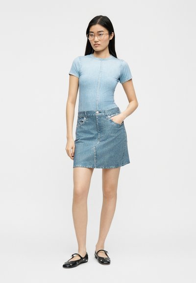 Γυναίκα με μαύρα μακριά μαλλιά και γυαλιά, που φοράει ένα γαλάζιο εφαρμοστό κοντομάνικο τοπ, τζιν φούστα και μαύρες μπαλαρίνες με λουράκια στον αστράγαλο.