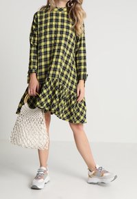 Abito a maniche lunghe a scacchi giallo e nero con orlo arricciato, abbinato a una borsa intrecciata bianca e sneakers grigie e bianche.