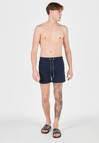 Pantaloni da bagno blu marina con vita elasticizzata e cordino verde. Presentano una texture liscia e arrivano fino a metà coscia.