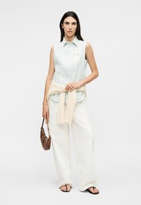 Femme portant une chemise bleu clair sans manches, un pantalon large blanc, un pull beige noué à la taille, des sandales noires, tenant un sac à main marron.