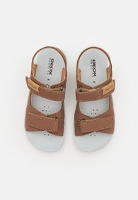 Sandalias de piel marrón con dos correas ajustables de Velcro, plantillas grises con perforaciones y un diseño de suela plana.