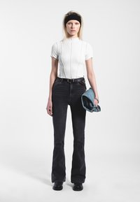 Valkoinen tiukka toppi, jossa on pystysuorat saumat, yhdistettynä mustiin leveälahkeisiin farkkuihin. Malli pitää kädessään rullattua farkkutakkia. Yksinkertainen vaalea tausta.