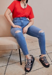 Rød t-shirt med korte ærmer, højttaljede revnede blå jeans og sorte sandaler med hvidt blomstermønster. Afslappet outfit på en beige stol.