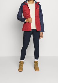 Röd och marinblå vattentät jacka med dragkedja framtill, kombinerad med marinblå leggings och bruna stövlar. Funktioner inkluderar huva och fack med dragkedja.