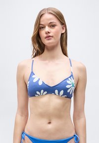 Νεαρή γυναίκα με μπλε μπικίνι τοπ με λευκά λουλούδια και μπλε μπικίνι σλιπ, μπροστά σε απλό φωτεινό φόντο.