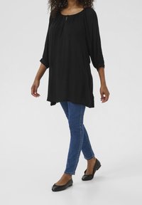Tunique noire avec un détail en forme de ключ-hole et des manches 3/4, associée à un jean skinny bleu et des ballerines noires, mettant en avant une coupe décontractée et une texture fluide.