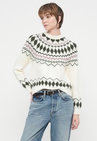 Ženska v belem puloverju z vzorcem, temno zelenimi in rožnatimi motivi, oblečena v svetlo modre kavbojke z črnim pasom, pozira na belem ozadju.