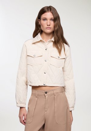 Giacca trapuntata cropped color crema con due tasche sul petto, bottoni a pressione e polsini elasticizzati, abbinata a pantaloni color marrone chiaro.