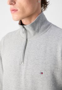 Pilkas zip-up džemperis su aukštu apykakle, pagamintas iš minkštos medžiagos. Savybės: sidabrinė užtrauktukas ir mažas siuvinėtas logo ant krūtinės.