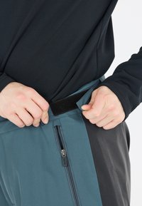Softshellbyxor i teal och svart, med en dragkedjeficka, justerbart kardborrebandsmidja och slät, elastisk tygkänsla.