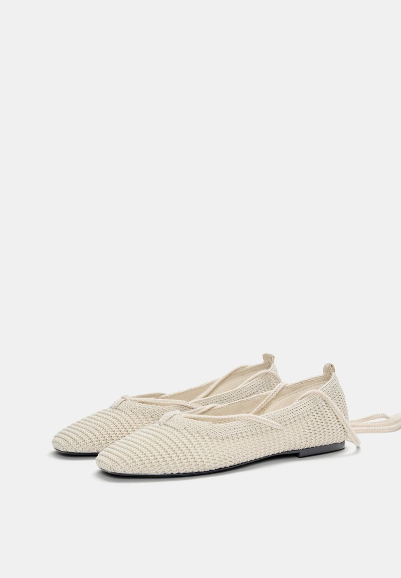 Ballerines tricotées beiges avec un motif texturé, un orteil arrondi et une semelle noire flexible. Comprend des lacets ajustables sur les côtés.
