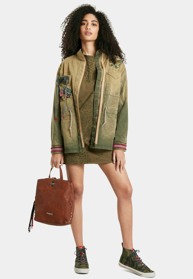 Giacca khaki multi-texturizzata con toppe ricamate, vestito verde a fantasia, zaino marrone a fantasia e sneakers alte mimetiche.