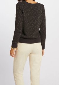 Femme portant un pull à imprimé léopard foncé et un pantalon blanc cassé, vue de dos avec un bracelet en or à son poignet.