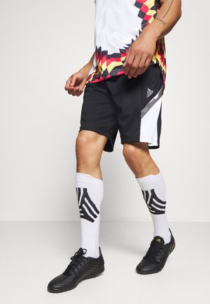 Person, die schwarze Adidas-Shorts, weiße Sportsocken mit schwarzem Design, schwarze Fußballschuhe und ein weißes Hemd mit einem roten, gelben und schwarzen geometrischen Muster trägt.