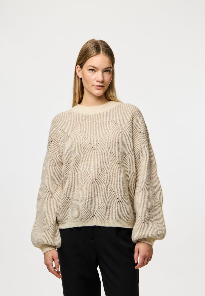 Beige strikket genser med et teksturert diamantmønster, vide ermer og rund hals. Bært med svarte bukser.