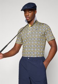 Polo s límečkem a krátkým rukávem v žluté, modré a bílé geometrickém vzoru; doplněno modrými šortkami. Model drží golfovou hůl.