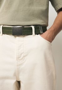 Cintura verde intrecciata con fibbia rettangolare in argento, indossata sopra pantaloni color crema con tasca laterale e dettagli di cuciture sottili.