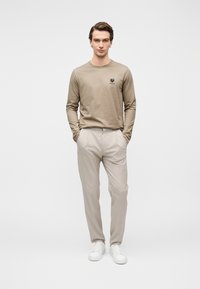 Μπεζ πουκάμισο μακρυμάνικο με μικρό λογότυπο, συνδυασμένο με ανοιχτόχρωμο παντελόνι με σχέδιο και λευκά αθλητικά παπούτσια, στέκεται μπροστά σε γκρίζο φόντο.