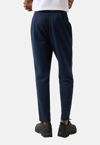 Osoba w granatowych spodniach dresowych i czarnych butach sportowych, stojąca z rękami skrzyżowanymi za plecami.