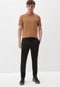 Bruine t-shirt met korte mouwen, gecombineerd met zwarte joggers met een trekkoord in de taille, gedragen met zwarte sneakers met tanzolen.