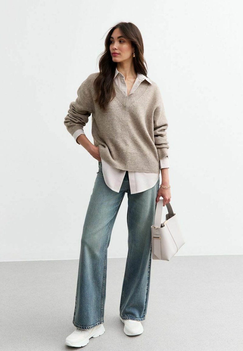 Beige oversized trui, witte blouse met kraag, lichtblauwe wijde spijkerbroek en witte sneakers. Houdt een kleine lichtgrijze handtas vast.