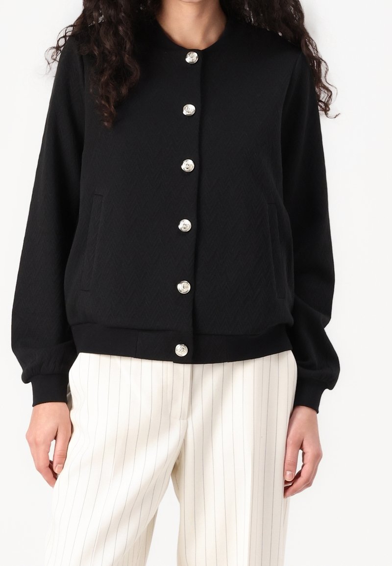 Femme portant un cardigan noir boutonné avec des boutons argentés et un pantalon blanc à fines rayures verticales, debout devant un fond uni.