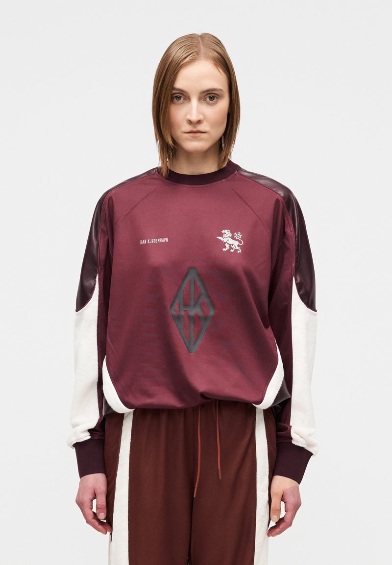 Sweat-shirt de sport bordeaux avec des manches contrastantes blanches et bordeaux foncé. Présente un logo de lion et un motif géométrique noir sur le devant.