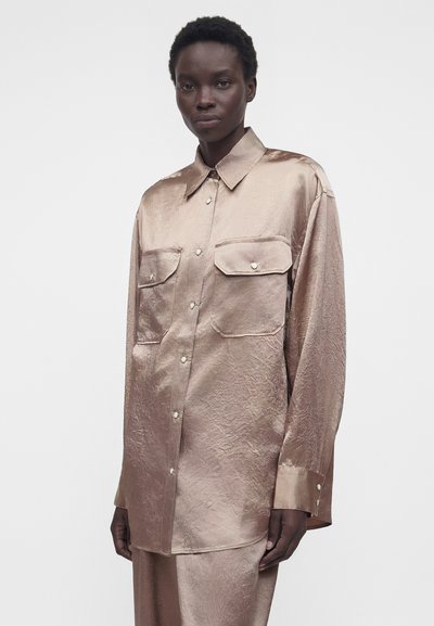 Άτομο σκούρου δέρματος που φορά χαλαρό, γυαλιστερό, ανοιχτό καφέ μακρυμάνικο πουκάμισο με κουμπωμένες τσέπες στο στήθος και ταιριαστό παντελόνι σε λευκό φόντο.